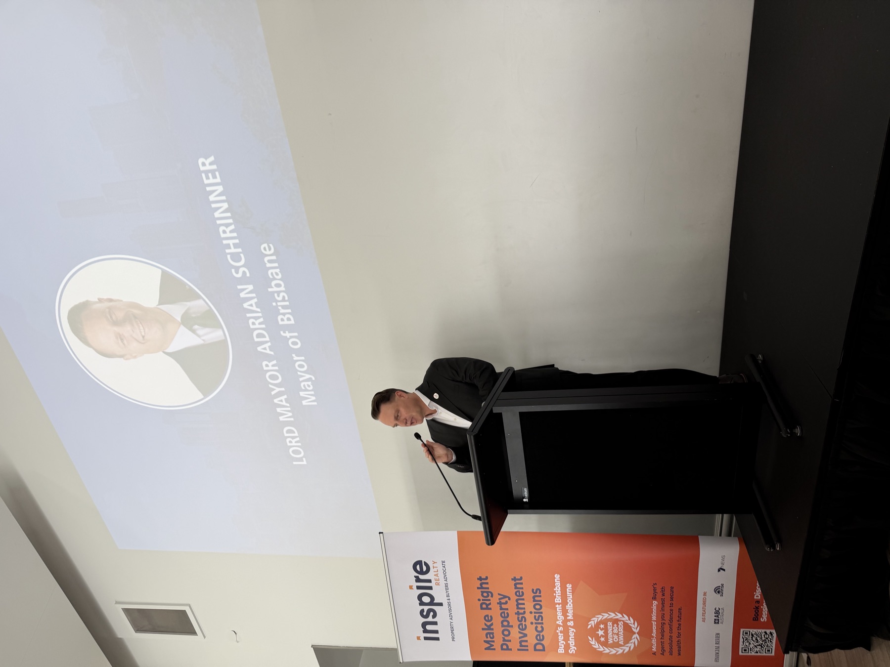 The Lord Mayor speaking at a lectern beneath a projected slide identifying Adrian Schrinner, Lord Mayor of Brisbane
