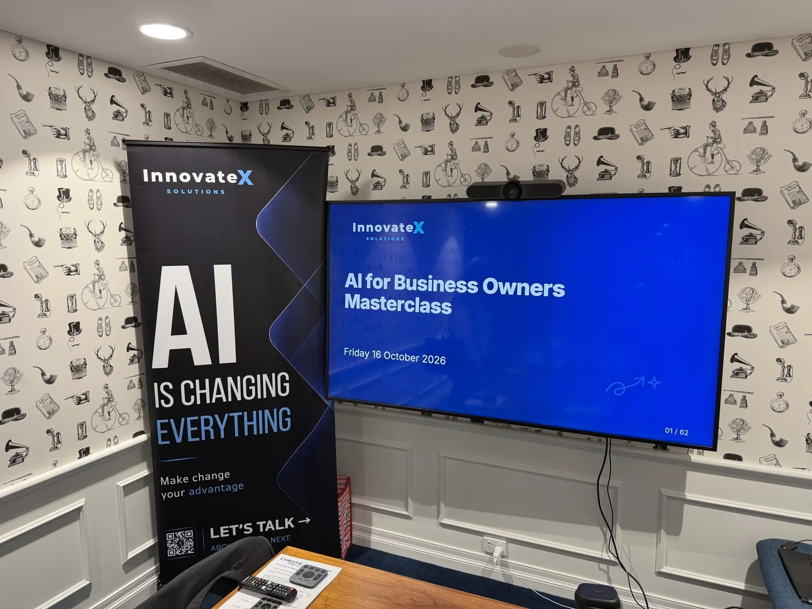 A meeting room with an InnovateX banner reading AI is changing everything beside a screen showing the AI for Business Owners Masterclass title slide